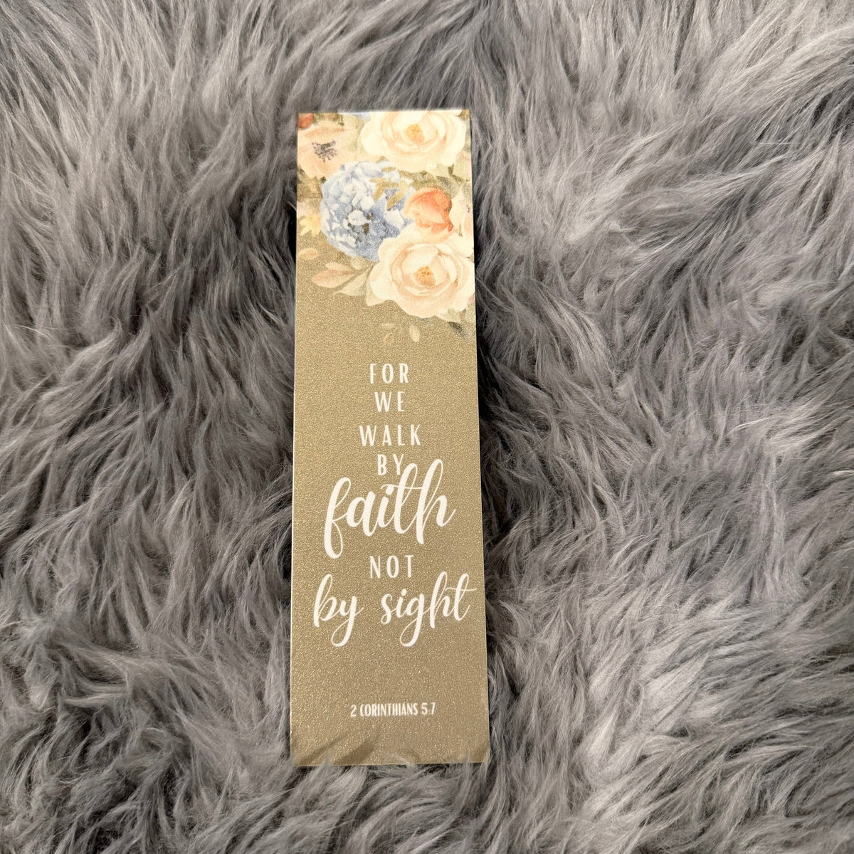 A rectangular decorative book or journal cover with a floral illustration at the top and the phrase 'FOR WE WALK BY faith NOT by sight' on a textured taupe background, placed on a gray faux-fur surface.